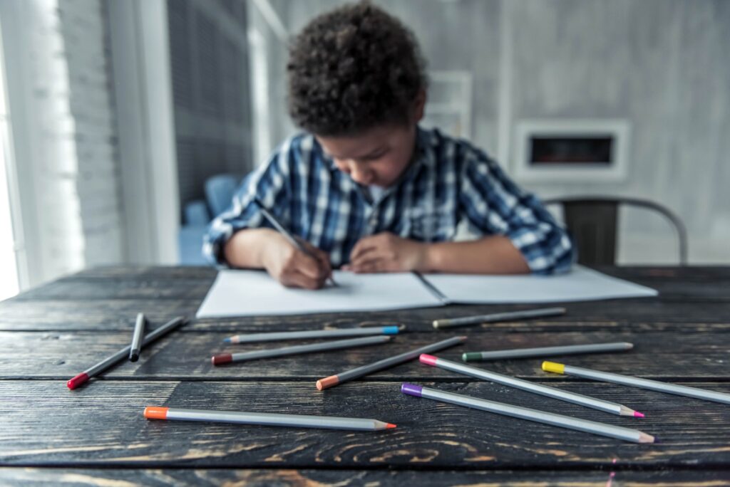 A child draws in a notebook at a table with multiple colored pencils.