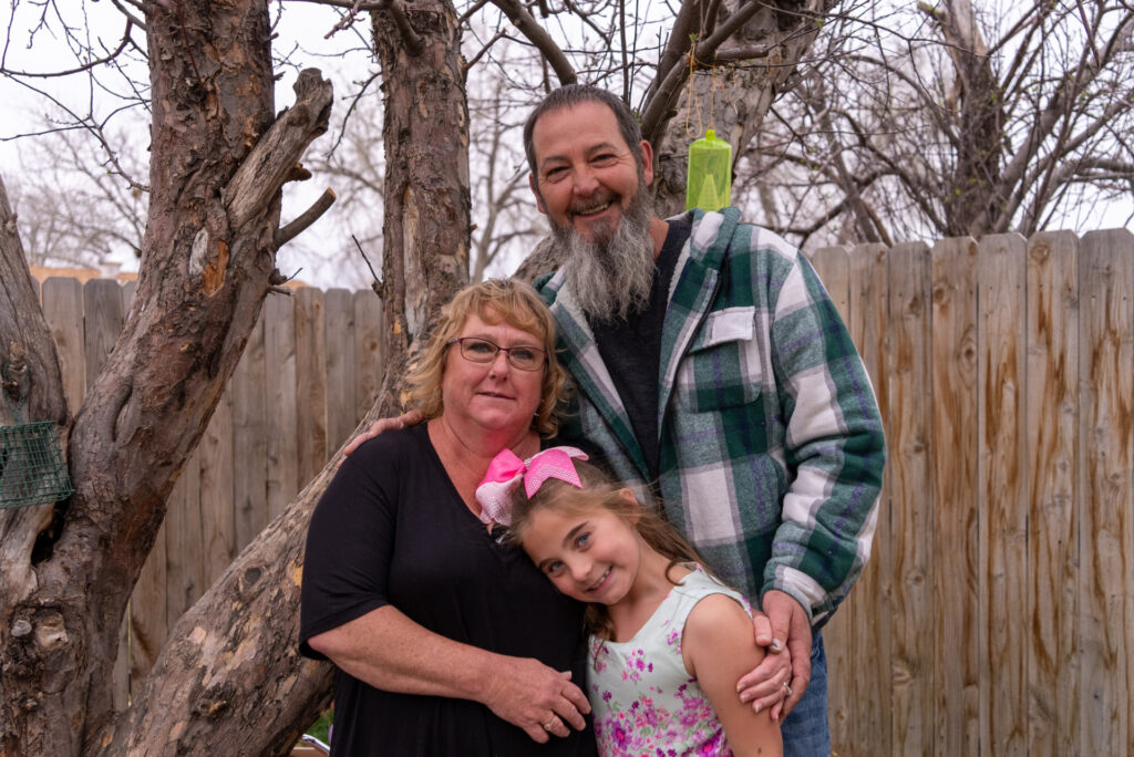 A kinship foster family of a father, mother, and daughter smile in their backyard.