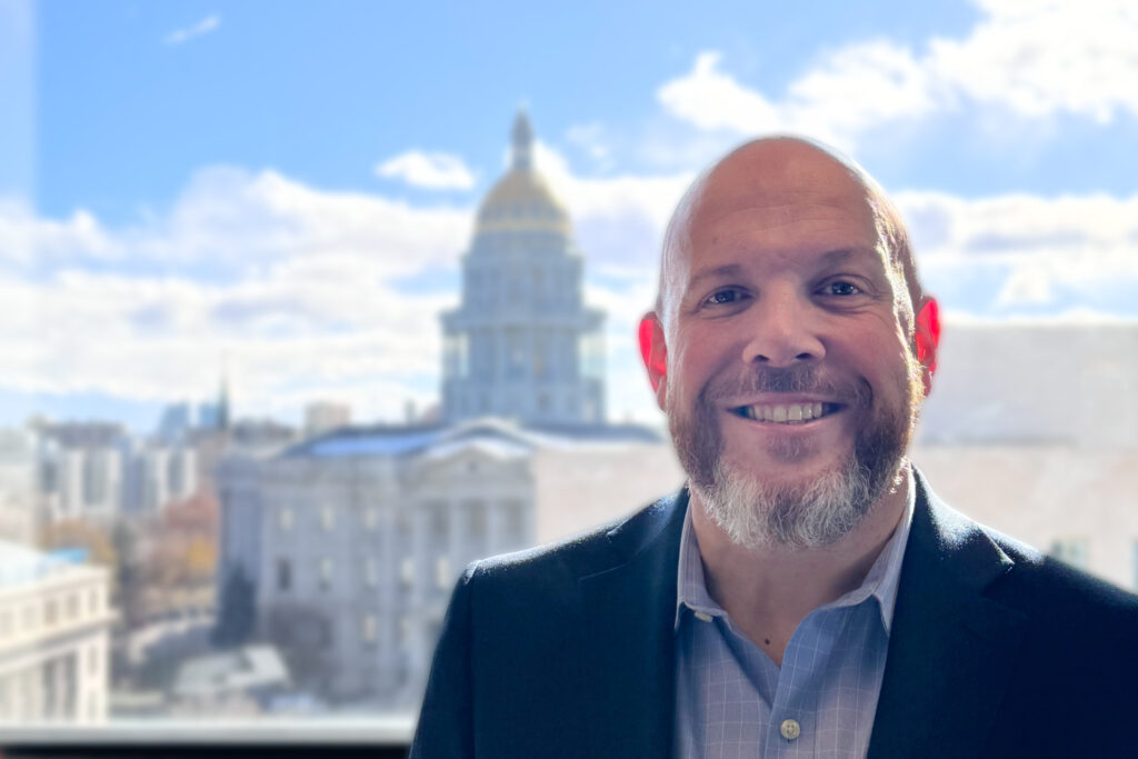 Smiling bearded man in professional dress stands on a roof with the Colorado State Capitol in the background.