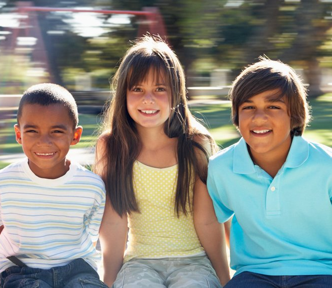 Tres niños sentados en un parque infanti, sonriendo a la cámara en un día soleado.