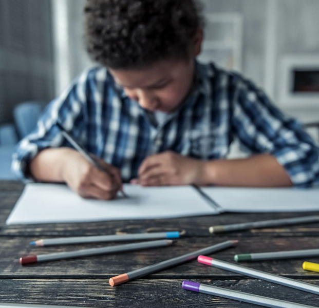 Un niño dibujando en un cuaderno en una mesa con varios lápices de colores.