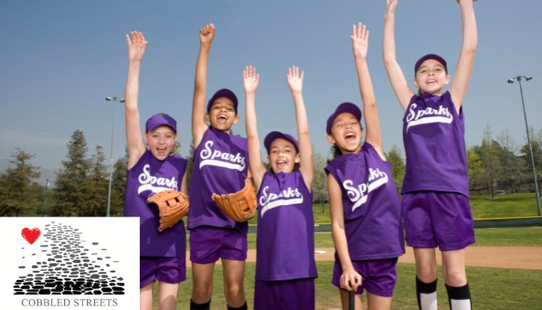 Young girls in matching uniforms celebrating on the softball field with the Cobbled Streets logo in the corner.