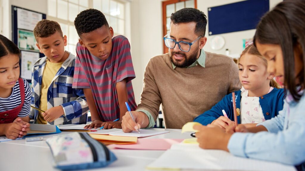 A male teacher writing on a sheet and five students around him paying attention