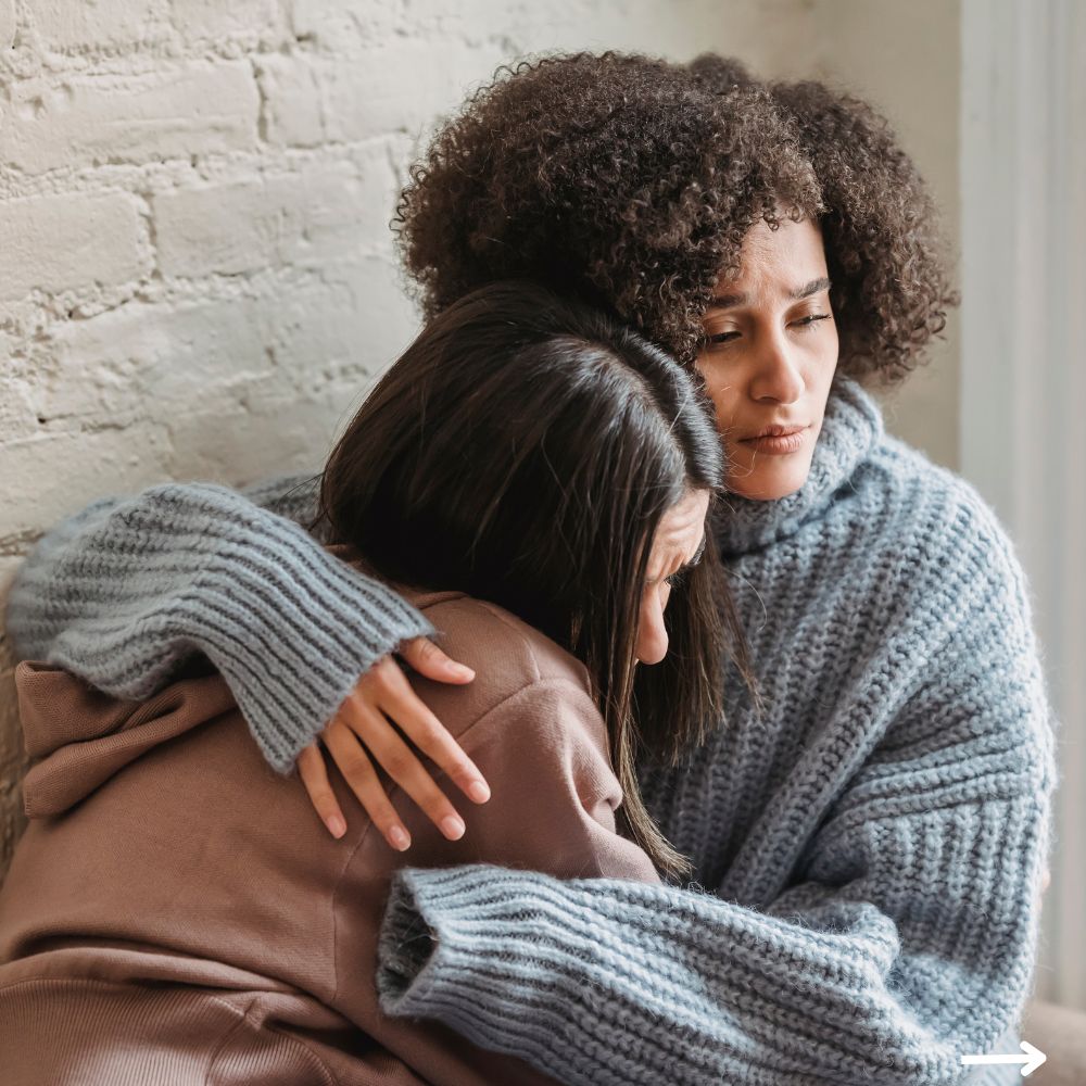 Two women, one with curly hair and a gray sweater, comfort each other while sitting against a white brick wall.