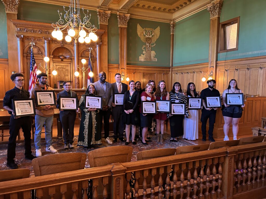 A group of young adults and adults, standing in rows, smile as they hold awards in a grand hall with wooden walls and chandeliers.