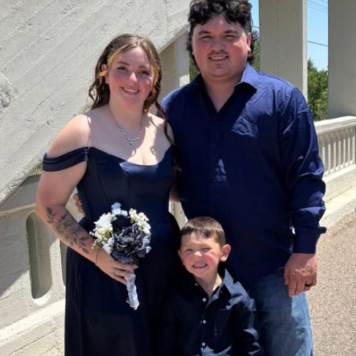 A family of three stands on a bridge, posing for a picture. The woman, in a dark blue dress, holds a bouquet of white and black flowers.