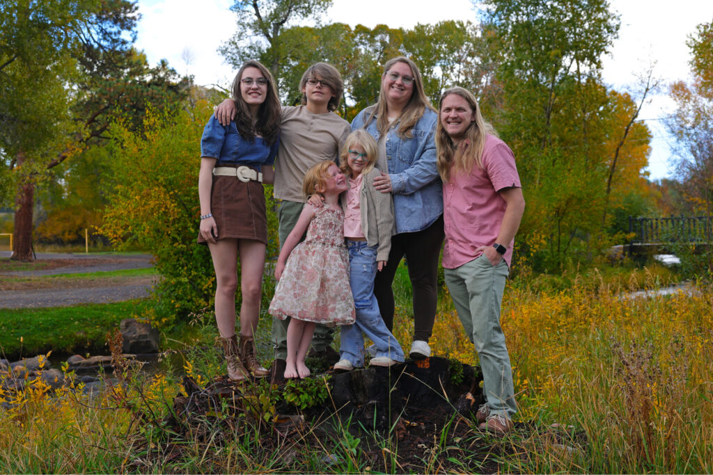 A family of six, two adults and four children, poses outdoors in front of trees with autumn foliage.
