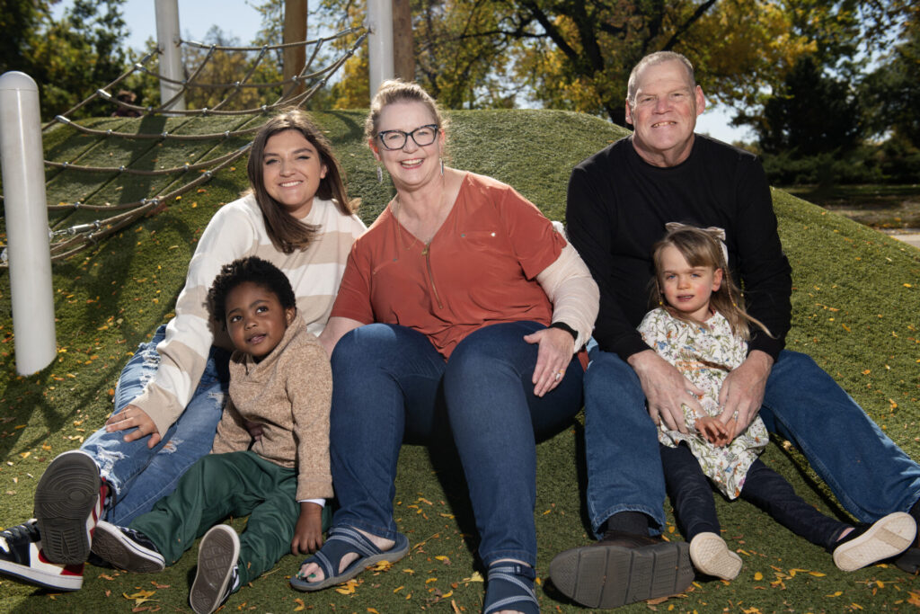 A family of five, including two adults and an adult child and two young children, sitting together on a grassy mound at a playground on a sunny day.