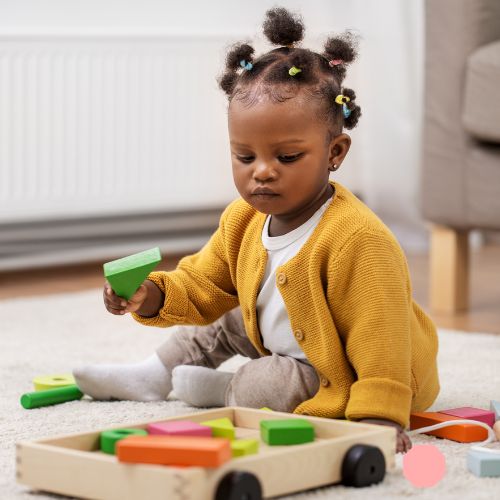 Toddler siting on a carpet playing with wooden blocks.