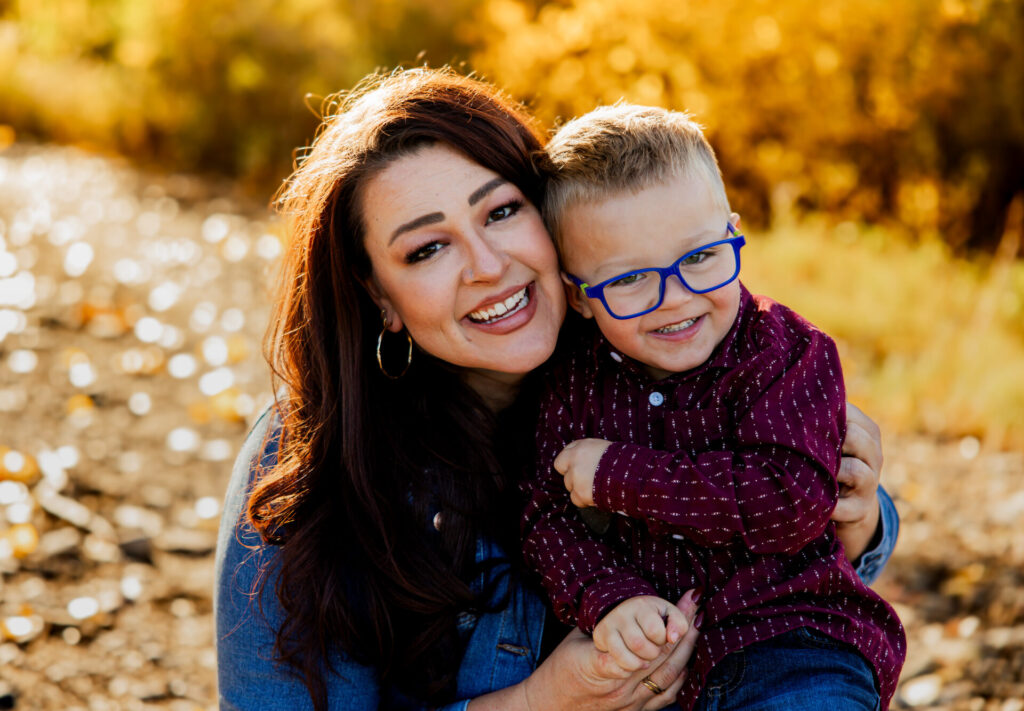 A smiling woman with dark hair wearing a denim jacket is holding a young boy wearing a burgundy shirt and blue glasses in an outdoor setting with a fall-colored background.