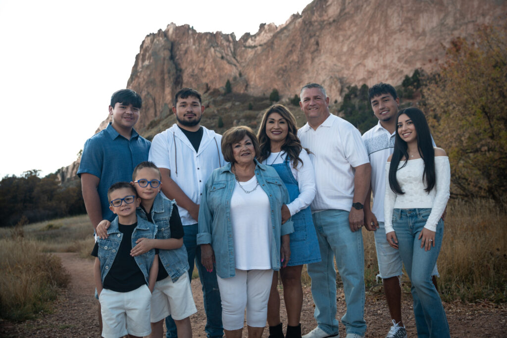 An outdoor family portrait features eight people, including two young boys and six adults, posing and smiling on a dirt path with a large, jagged reddish-brown mountain in the background.