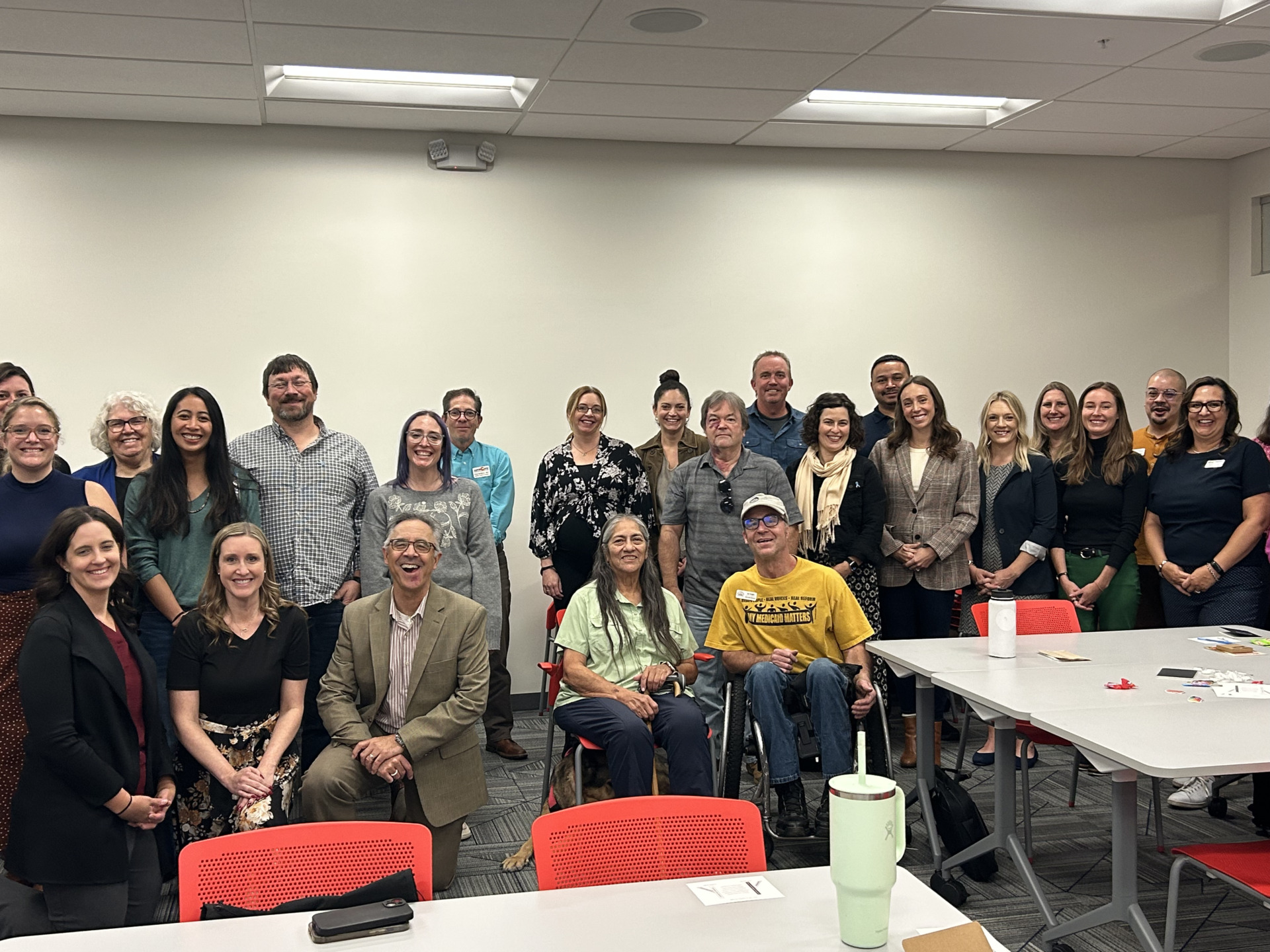A diverse group of people is posing together for a photo in a brightly lit room with white walls, some standing and some seated, including one person in wheelchair.