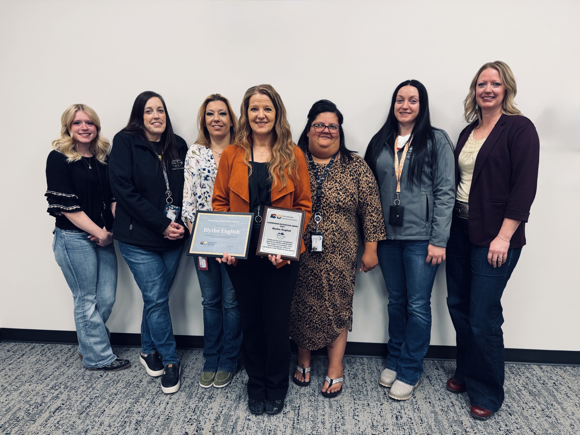 The photo shows seven women standing side-by-side in a room with white walls and gray carpet, with one woman in the center holding two award plaques that bear the name "Blythe English".