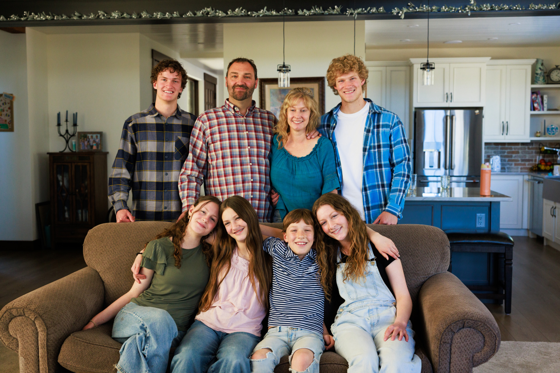 A large family with a mother, father, and six children—three girls and three boys—pose together in their living room, with the four adults and two older boys standing behind four younger children seated on a brown sofa.