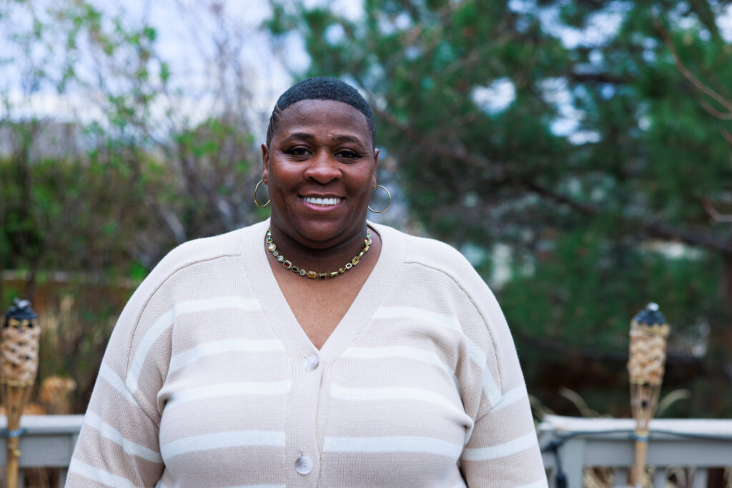 A Black woman with a short haircut, wearing a beige and white striped cardigan and a gold-colored choker necklace, smiles while standing outside with a blurred background of greenery.