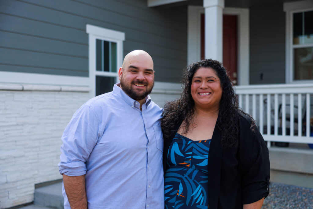 A smiling couple, a man in a light blue collared shirt and a woman in a patterned dress and black cardigan, stand outdoors in front of a gray house with white trim.
