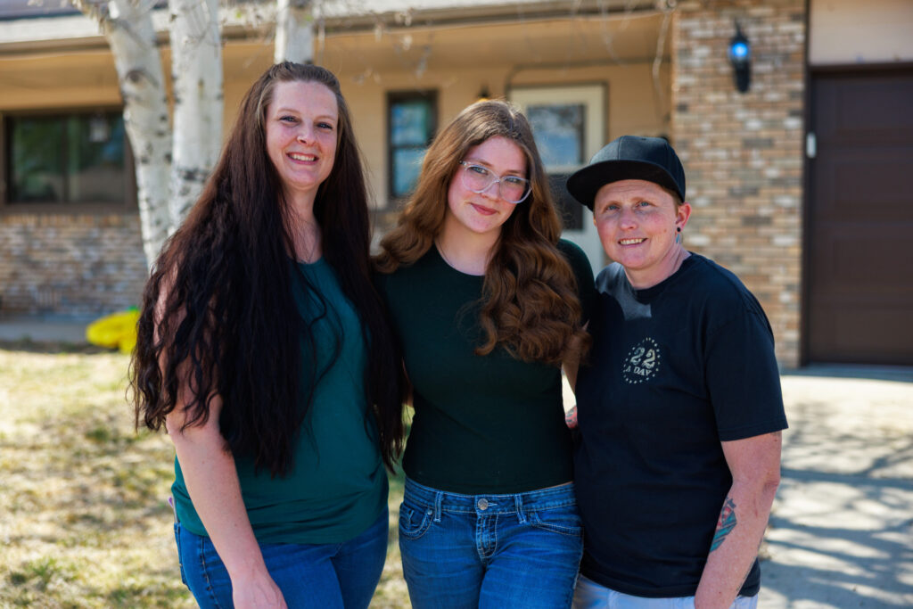 Two moms pose with their teenage daughter in font of their house.
