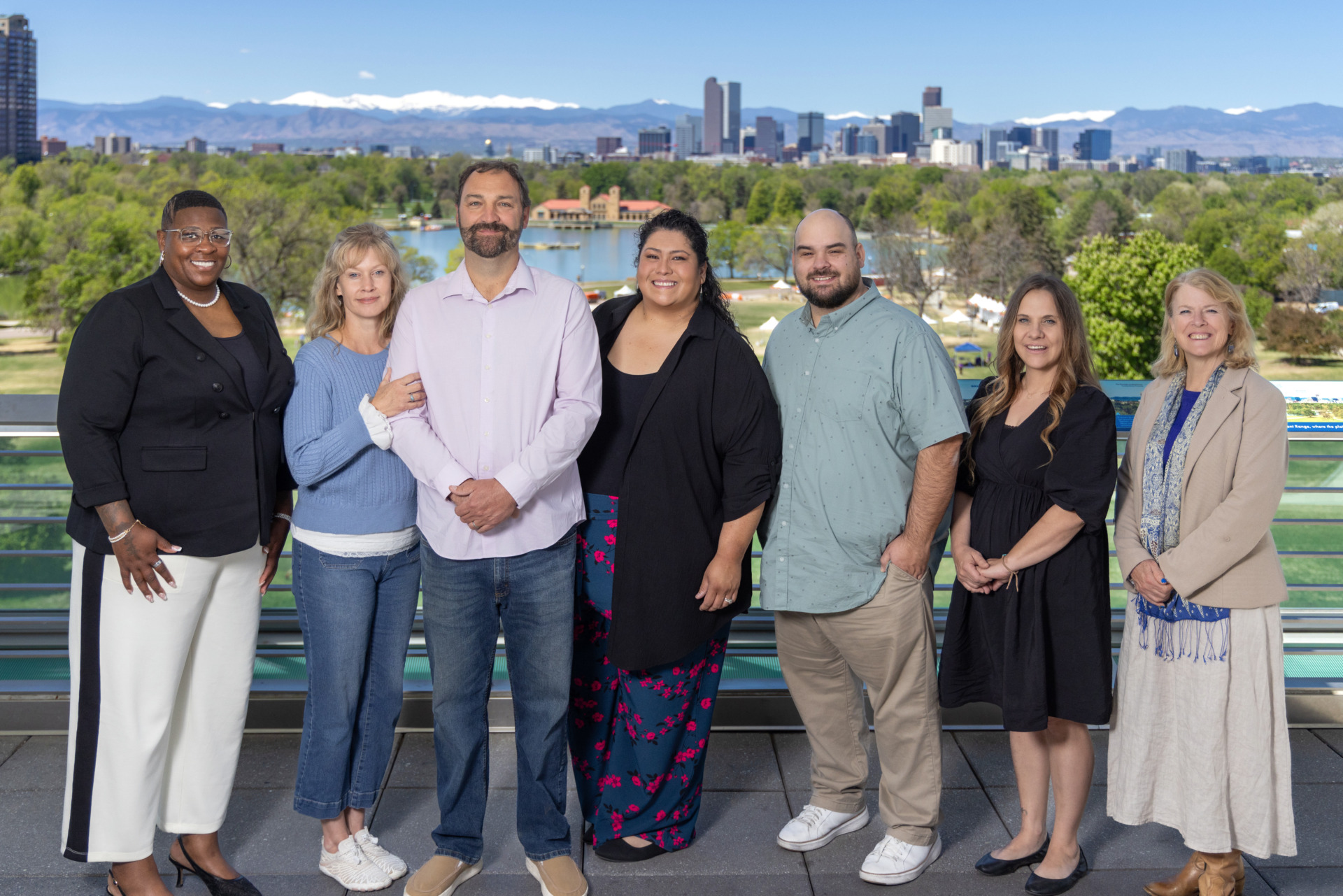 A diverse group of five women and two men pose for a photo on a balcony overlooking a park and downtown Denver.