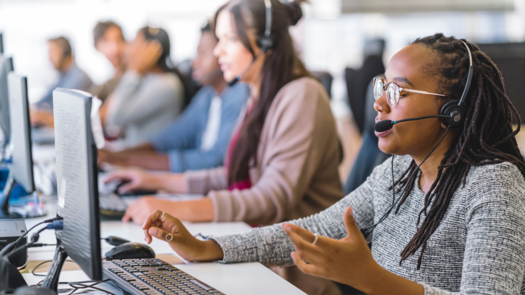 An African American female call taker responds to an incoming call in a bustling call center, surrounded by colleagues answering phones in the background.