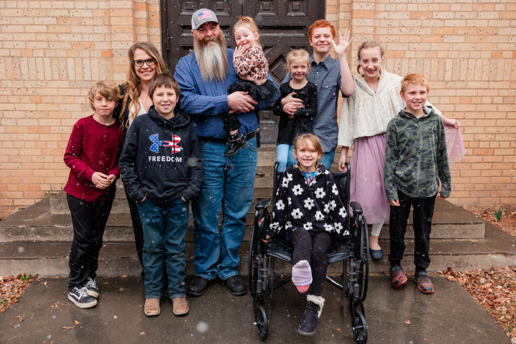 Family with mom and dad and eight children stand in front of a brick building in the snow.