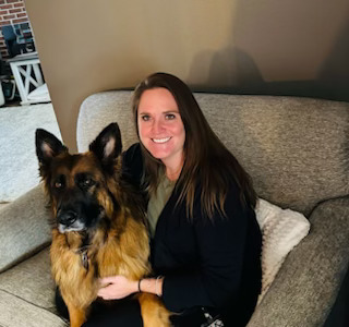 A woman with long brown hair smiles while sitting on a couch next to a large German Shepard dog.