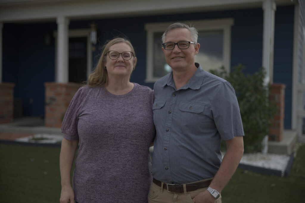A middle-aged couple in glasses smiles together outdoors in front of a navy blue house with white trim. The man wears a grey short-sleeve shirt.