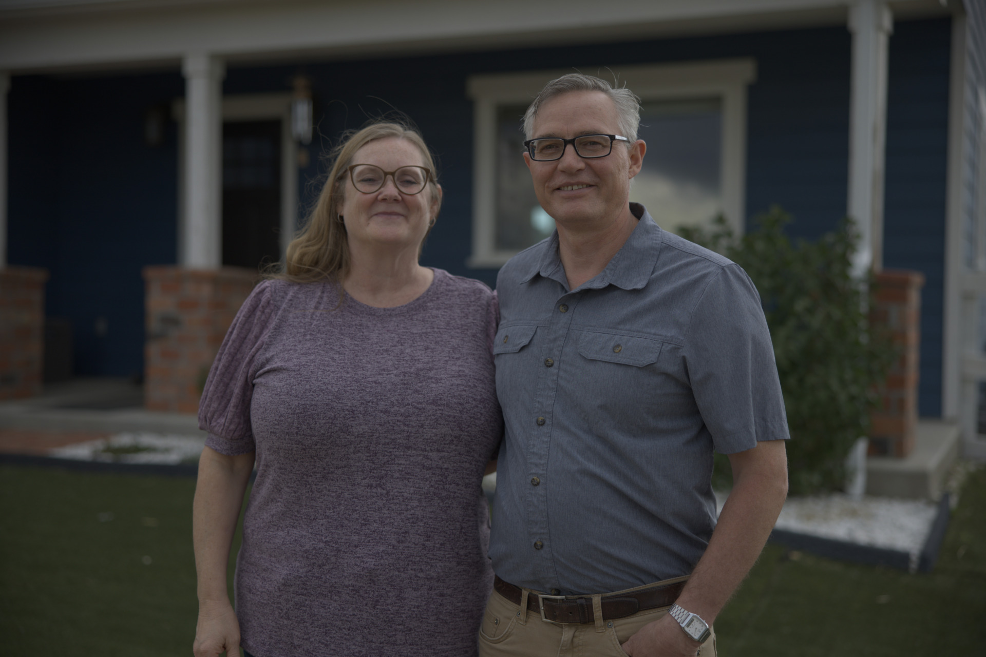 A middle-aged couple in glasses smiles together outdoors in front of a navy blue house with white trim. The man wears a grey short-sleeve shirt.