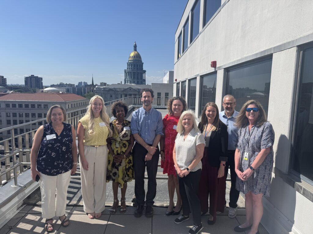 A group of nine people poses outside on a sunny rooftop terrace, with a gold-domed state capitol building visible against a clear sky in the distance.