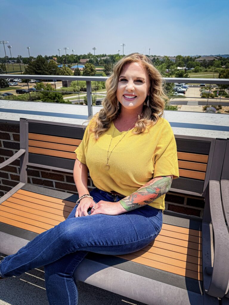 A woman with blonde hair, a yellow shirt, and a colorful arm tattoo smiles while sitting on a wooden bench outdoors on a sunny rooftop patio.