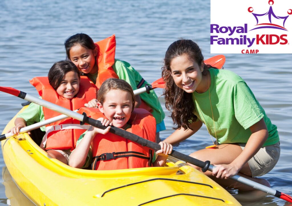 A counselor stands in water next to three young girls wearing life jackets in a yellow kayak. Top right corner reads "Royal Family KIDS CAMP."