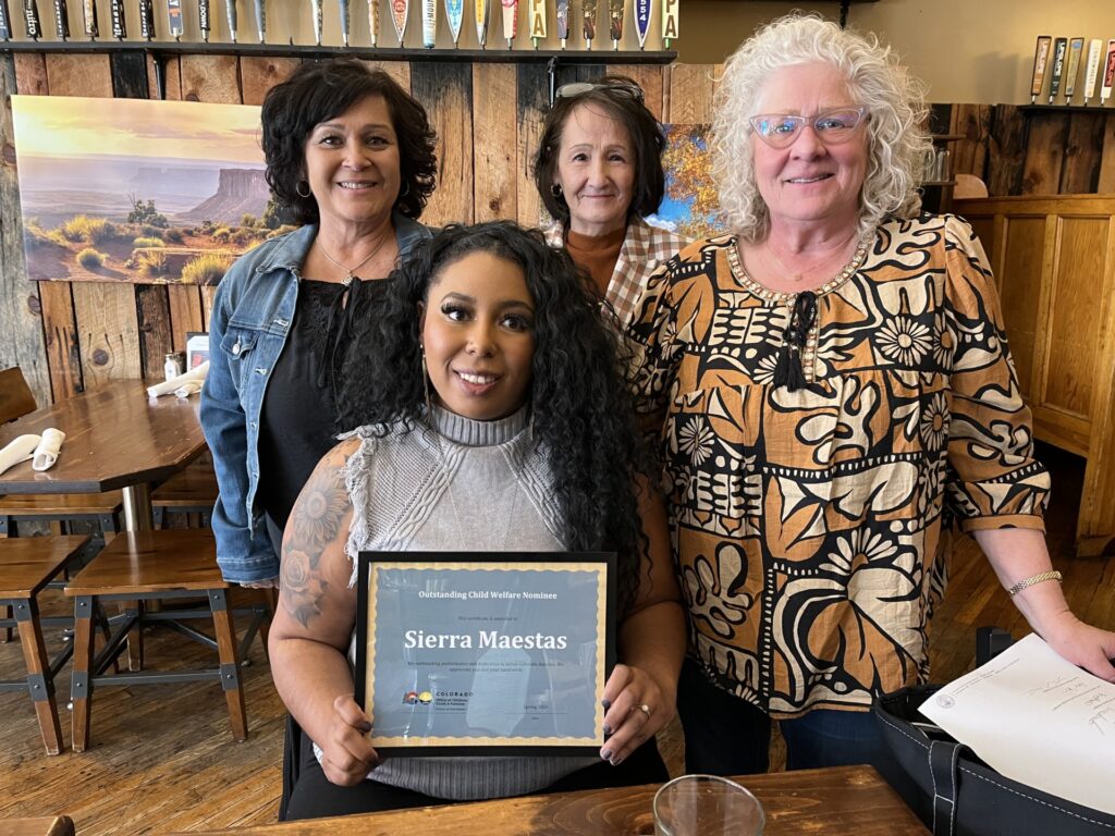 Sierra Maestas sits smiling, holding an Outstanding Child Welfare Nominee certificate, surrounded by three supportive women standing behind her.