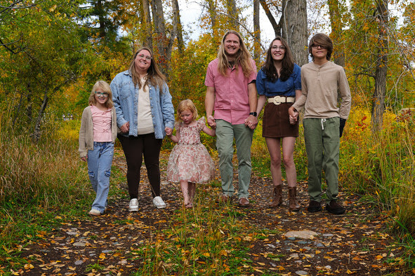 A smiling family of six holds hands while standing together on a leaf-strewn dirt path surrounded by colorful autumn trees.