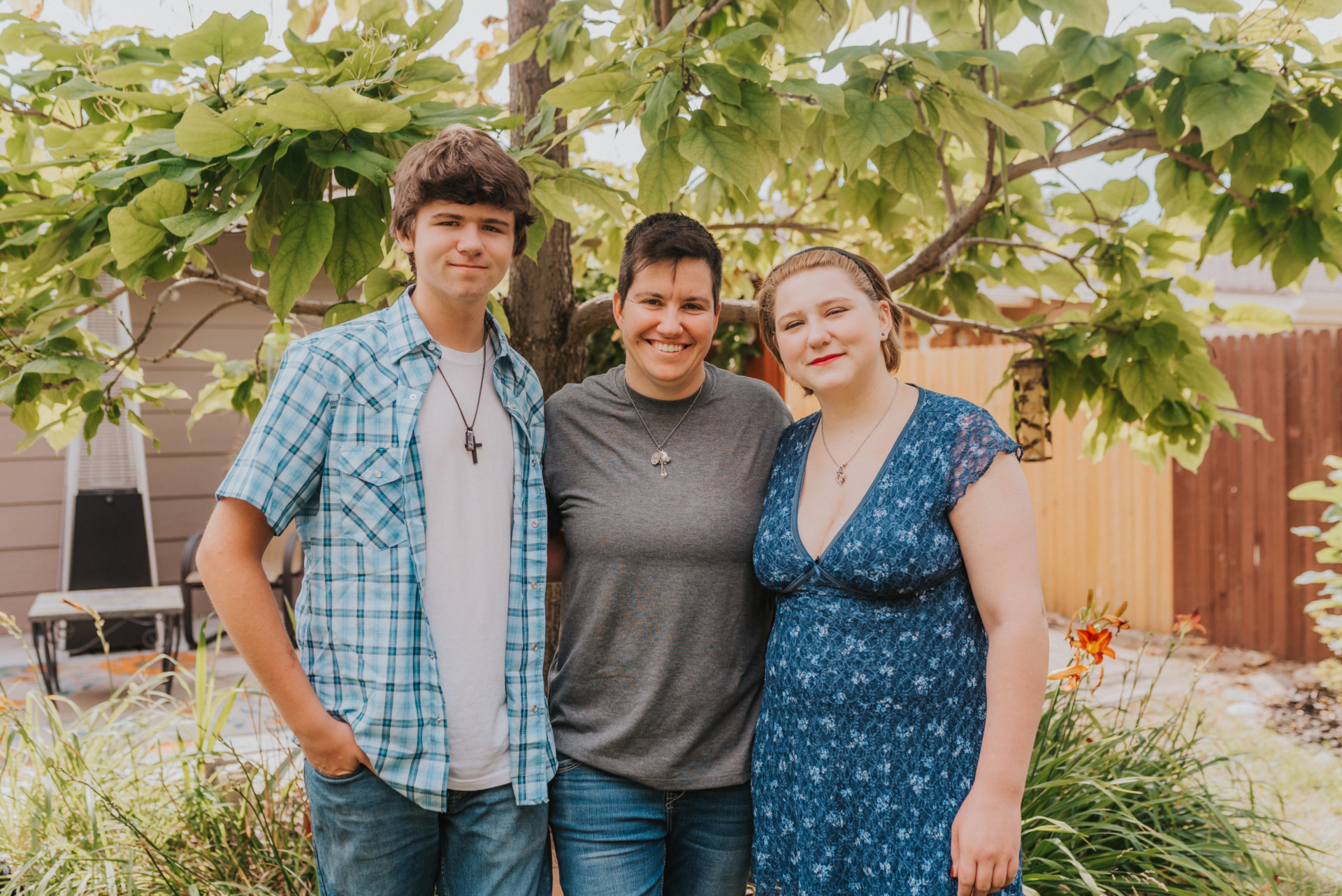 A smiling woman stands outdoors between a young man in a blue plaid shirt and a young woman in a blue floral dress under a leafy tree.